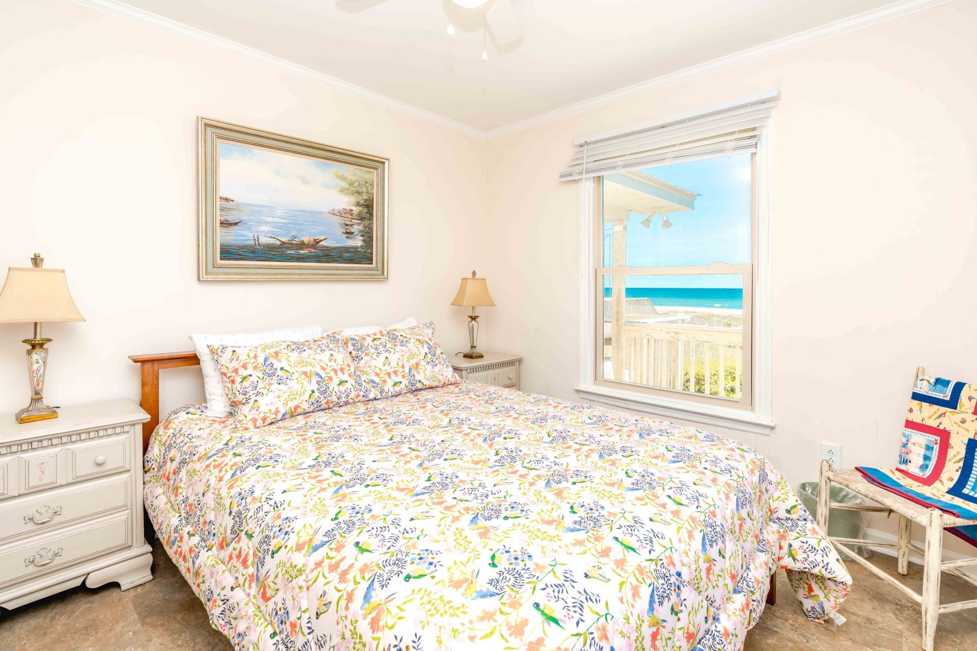 A bedroom with a bed, two bedside tables, a nightstand, a window with a view of the beach, and a painting on the wall.