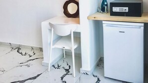 Een kleine kamer met een wit bureau, een witte stoel, een houten plank met een strijkijzer, en een witte koelkast.