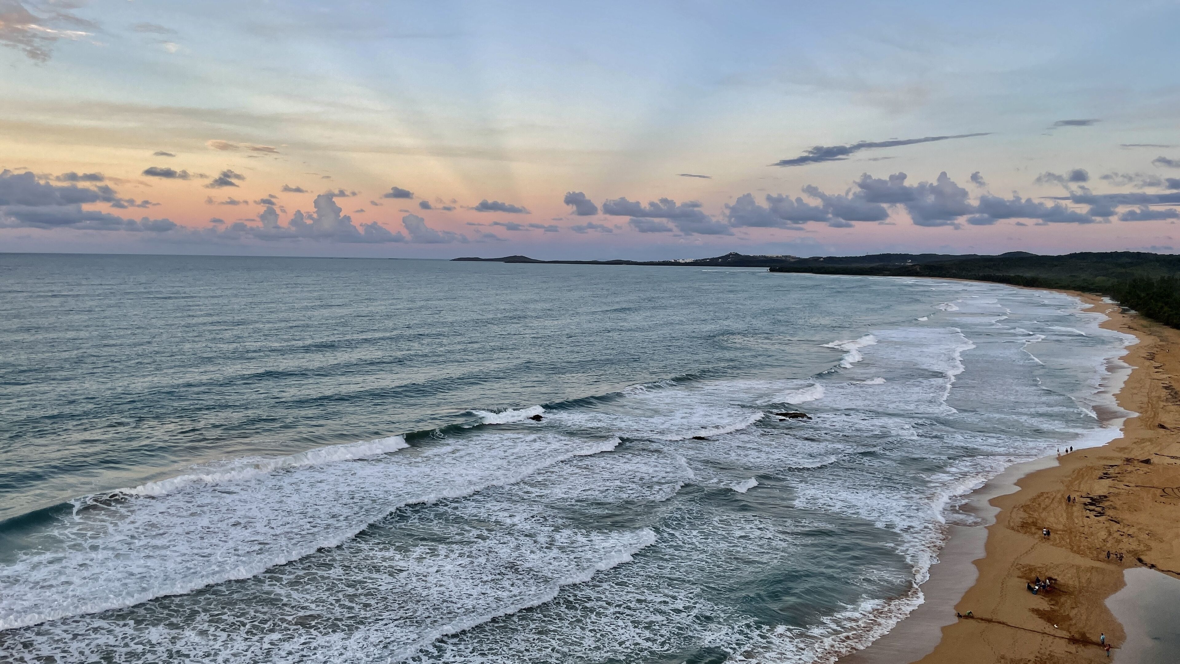 A beach with waves crashing onto the shore, a clear sky, and distant landforms.