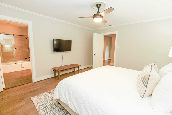 Un dormitorio con cama, ventilador de techo, televisor, banca y baño visible a través de una puerta abierta.