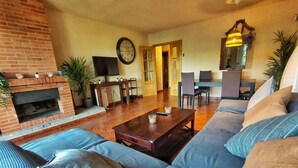A living room with a brick fireplace, a large sofa set, a wooden coffee table, and a dining area with chairs and a table. - Casa Rural La Goleta.  Pantano de San Juan (San Martín de Valdeiglesias)