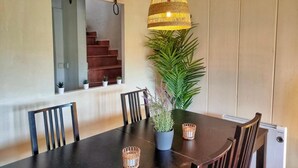 A dining area with a dark wooden table, four chairs, a hanging light fixture, a potted plant, and a staircase in the background. - Casa Rural La Goleta.  Pantano de San Juan (San Martín de Valdeiglesias)