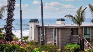 A beachfront building with a balcony overlooking the ocean, surrounded by palm trees and various plants. - Coastal One-Bedroom Haven with Ocean Peeks and Serene Gardens (Oceanside)