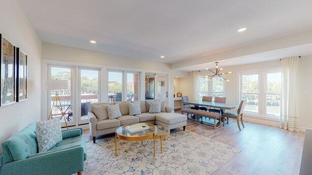 A spacious living room with a sectional sofa, a glass coffee table, and a dining area with a table and chairs. Large windows provide ample natural light and views of the outdoors. - Completely Updated Two Bedroom Condominium with Sweeping Marina Views! (Hilton Head)