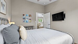 Un dormitorio con cama, almohadas, ventilador de techo, televisor y una pared decorada con cuadros.