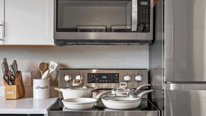 Una cocina con microondas, horno y refrigerador de acero inoxidable. Hay ollas en la estufa, un organizador de utensilios y un estante para especias.