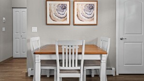 Comedor con mesa de madera y sillas blancas, dos cuadros enmarcados sobre la mesa y una puerta blanca a la derecha.