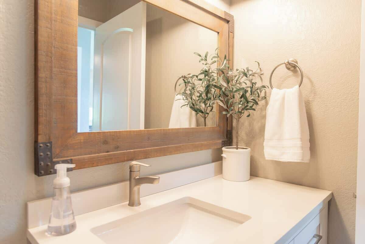 A bathroom with a wooden-framed mirror, a white sink, a potted plant, a towel ring, and a soap dispenser.