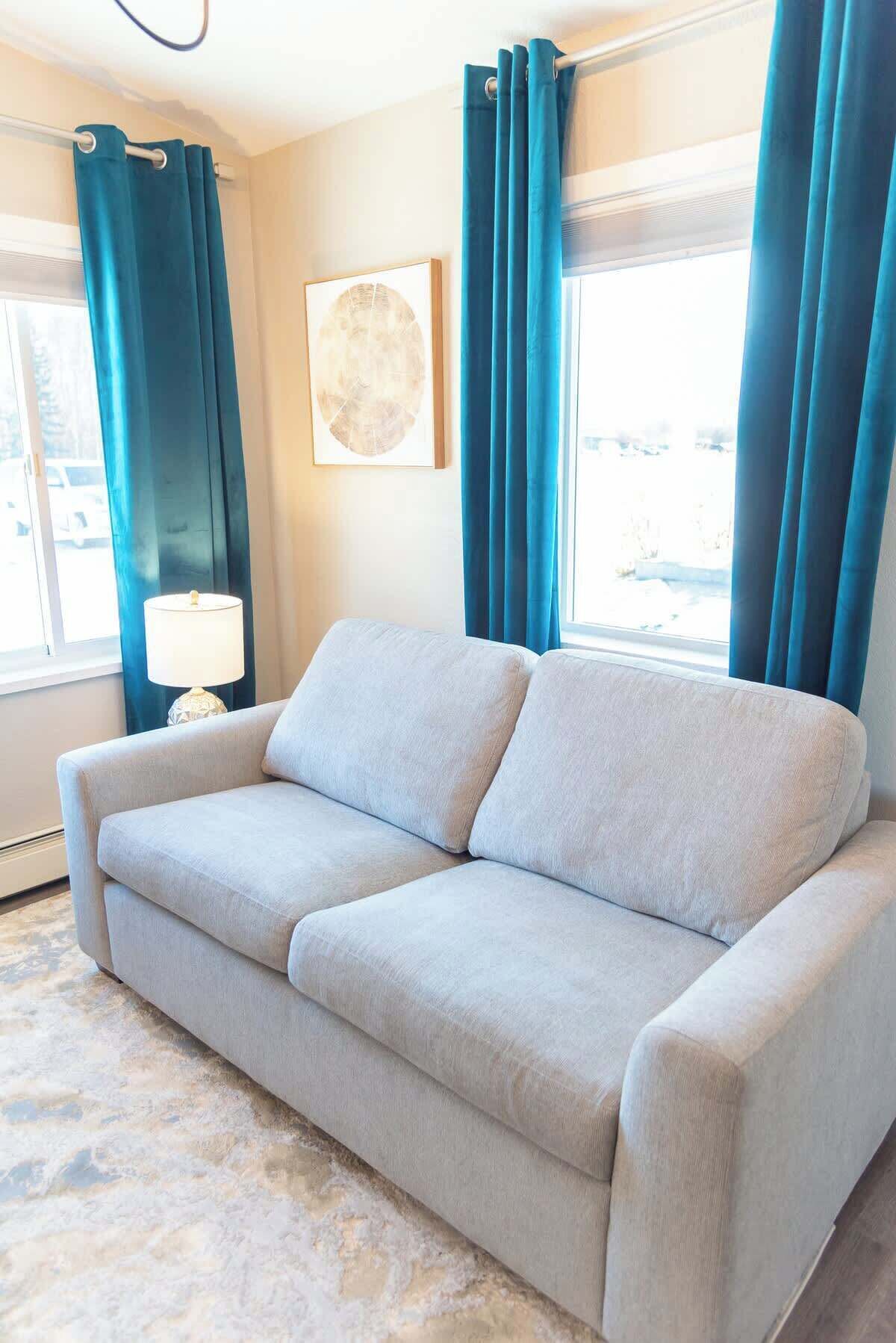 A light grey sofa in a room with a large window, blue curtains, and a framed artwork on the wall.