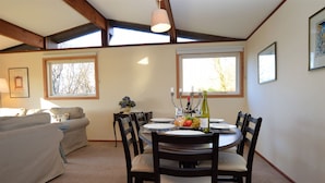 Un coin repas avec une table ronde dressée pour quatre personnes, entourée de chaises en bois. Une fenêtre donne sur un paysage arboré, et un canapé est visible au fond.
