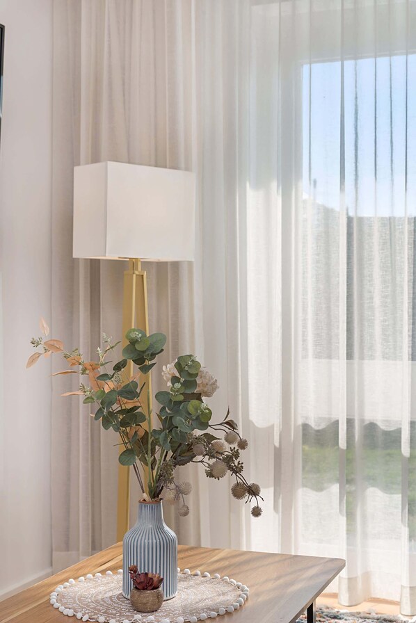 A room with a wooden table, a blue vase with plants, a white lamp, and sheer curtains.