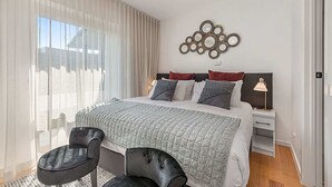 A modern bedroom with a large bed, two chairs, a bedside table with a lamp, and a decorative wall piece. - Comfy Studio with Shared Backyard near Racecourse (Carnegie)