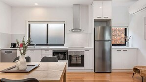 A modern kitchen with white cabinetry, a stainless steel refrigerator, a built-in oven, and a window with a brick wall view. - Gorgeous 3-Bed House with Spacious Shared Backyard (Carnegie)