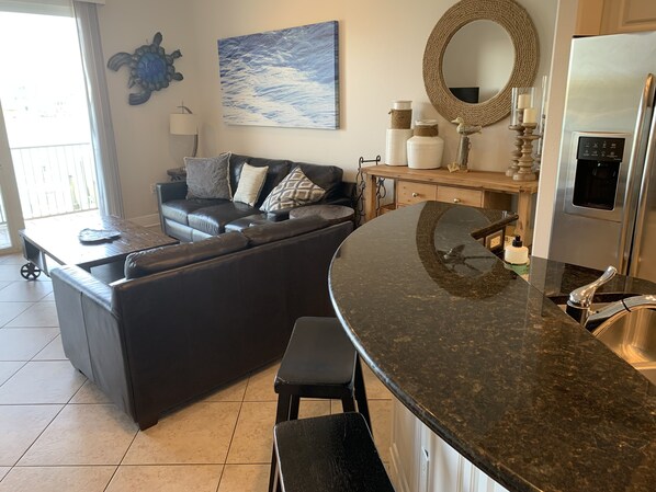 A modern kitchen with a curved granite countertop, a stainless steel refrigerator, and a dining area with a dark leather sofa and patterned cushions.