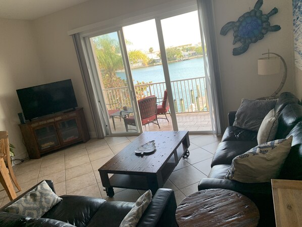 A living room with a sofa, a coffee table, and a TV on a cabinet. There is a sliding glass door leading to a balcony with a view of a body of water and a building. - Luxury Spacious Getaway Perfect for Larger Families or Groups (Clearwater)