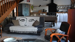 A living room with a sofa, a wooden chair, a dining table, and a kitchen area with a stove and cabinets. - La Maison d'Anne-Marie, charming vacation rental (Saint-Clet)