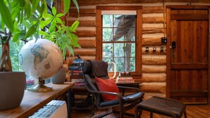 Um quarto de cabana rústica e acolhedor, com uma escrivaninha de madeira, um globo, uma cadeira com almofada vermelha, uma janela com vista para árvores e um tabuleiro de xadrez.