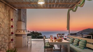 A terrace with a table set for dining, a sofa, and a view of the sea at sunset. - Arca Villa   Enchanting Sunset! (Keri)