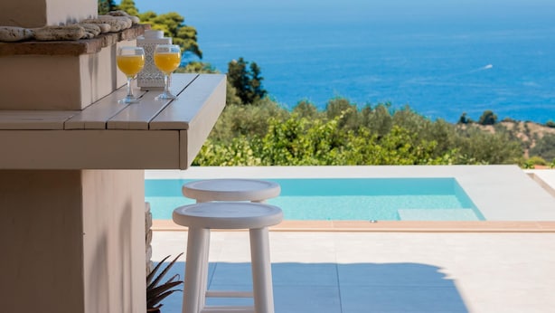 A poolside area with a view of the ocean, two glasses of orange juice, and a stone bench. - Arca Villa   Enchanting Sunset! (Keri)