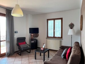 Ein Wohnzimmer mit einer Couch, einem Sessel, einem Couchtisch, einem Fernseher, einem Fenster und einer Lampe.