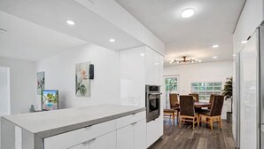 A modern kitchen with white cabinetry, a built-in oven, and a dining area with wooden chairs and a glass table. - Luxe Mansion/10min Walk Beach/Gameroom/Heated pool (Hollywood)
