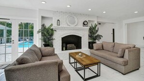 A modern living room with a white brick fireplace, two sofas, a coffee table, and a pool visible through glass doors. - Luxe Mansion/10min Walk Beach/Gameroom/Heated pool (Hollywood)