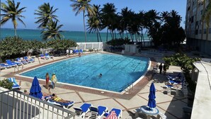 A swimming pool with lounge chairs and umbrellas, surrounded by palm trees and a view of the ocean. - Oceanfront on 17th Floor in Condado (San Juan)