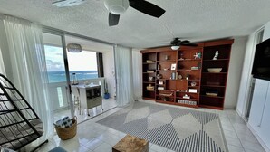 A spacious living room with a large bookshelf, a blue sofa, a wooden coffee table, and a view of the ocean through large windows. - Oceanfront on 17th Floor in Condado (San Juan)