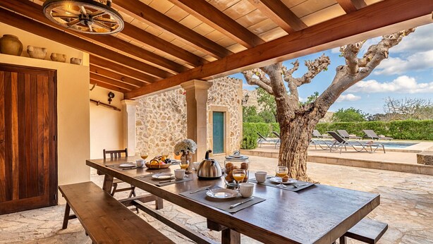 Un espacio al aire libre con comedor, una mesa de piedra dispuesta para el desayuno, techo de madera y vistas a una piscina y follaje.