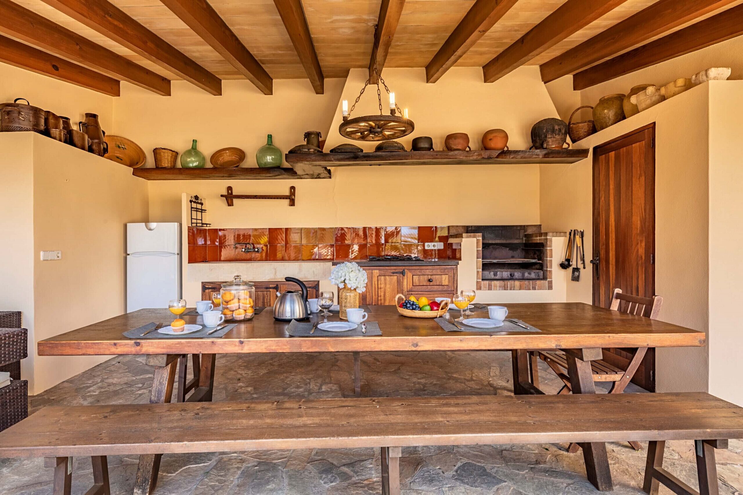 A rustic kitchen with wooden beams, a stone floor, and a dining table set with breakfast.