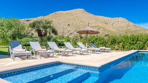 A swimming pool with lounge chairs and an umbrella, surrounded by greenery and a mountain in the background.