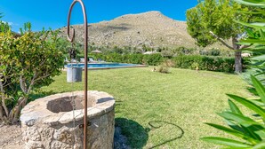 A stone well, a swimming pool, and a grassy yard with trees and a hill in the background.