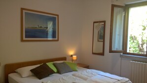 A bedroom with a bed, pillows, a nightstand with a lamp, a framed painting, and a window with a view of greenery. - Casa Maffei PLUS apartment 4, 1st floor, balcony, wonderful lake view, parking (Tignale)
