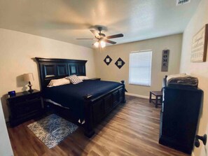 A bedroom with a bed, a nightstand, a dresser, a ceiling fan, a window with blinds, and a wall decoration. - The Ranch House near Medina Lake with SUNSET RESORT access! (Mico)