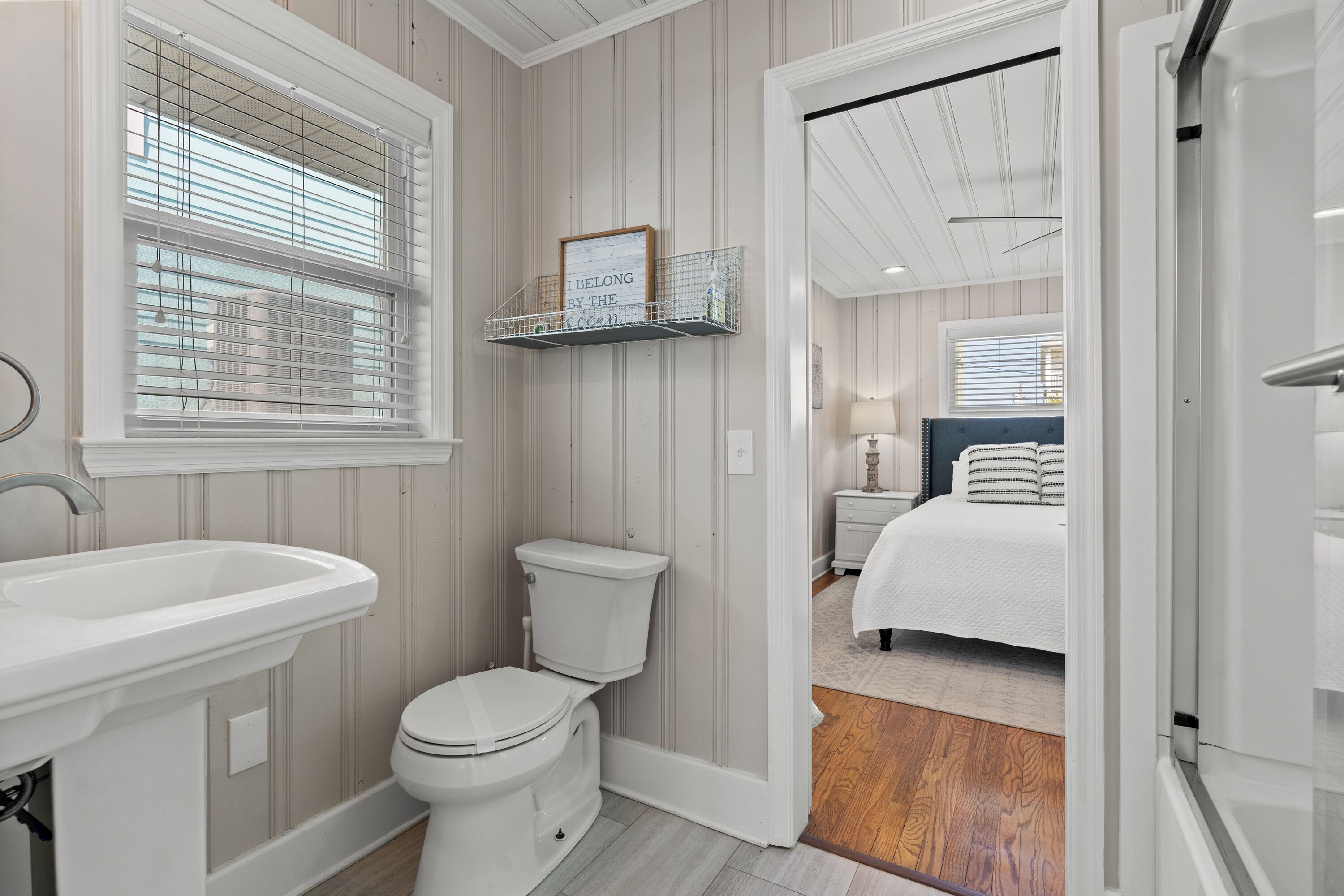 Une salle de bain avec un lavabo blanc, des toilettes et des stores aux fenêtres. Il y a une étagère avec un panneau encadré au-dessus des toilettes.