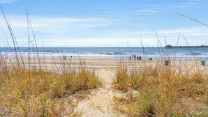 A beach with a pier in the background, sandy path leading to the water, and tall grasses in the foreground.