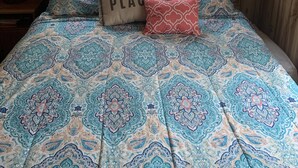 Une chambre avec une couvre-lit à motifs, un coussin orné de l’inscription « ICI, C’EST MON PETIT BONHEUR », et une fenêtre avec des stores.