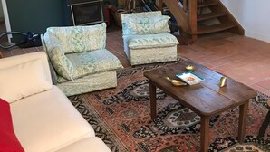 Una sala de estar con un sofá blanco, dos sillones estampados, una mesa de centro de madera y una alfombra con diseño. Una escalera de madera conduce a un nivel superior.