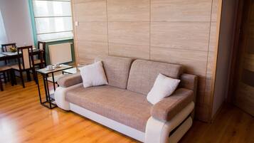 A modern living room with a beige sofa, two white cushions, a wooden floor, a dining table with chairs, and a large window with blinds.