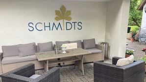 Un coin salon aménagé avec des meubles en osier, comprenant un canapé, deux fauteuils et une table. Un logo représentant une feuille d’érable et le texte « SCHMIDS » est affiché sur le mur.