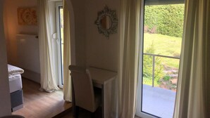 Une chambre comprenant un lit, un bureau, une chaise, un radiateur, une fenêtre avec des rideaux et donnant sur un jardin.