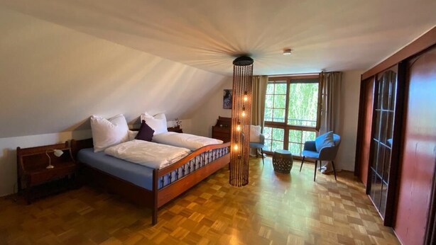 Une chambre avec un plancher en bois, un lit recouvert de draps blancs, un luminaire suspendu et une fenêtre avec des rideaux.