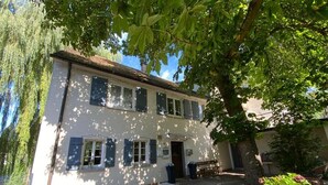 Une maison à deux étages avec une cour dallée, un banc et un arbre qui offre de l’ombre.