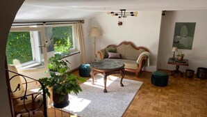 Un salon avec un parquet en bois, une table basse ronde, un canapé et un fauteuil. Il y a une fenêtre avec des rideaux, une lampe et un tableau au mur.