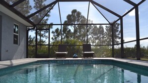 A swimming pool with two lounge chairs and a view of trees through a glass enclosure. - Brand new heated pool home. (Rotonda West)