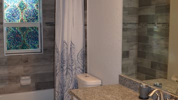 Un baño con lavamanos de encimera de granito, inodoro, ducha con puerta de vidrio y ventana de mosaico.