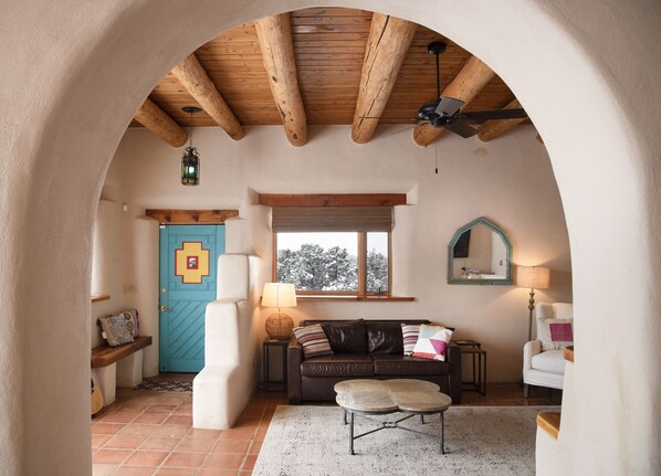 A cozy room with a wooden ceiling, a blue door, a window with a view of snow-covered trees, a brown leather sofa, a white armchair, and a small table with a lamp. - Peaceful Adobe home between Taos/TSV- Mountain Views, Hot Tub, Summer Garden (Valdez)