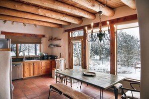 A rustic kitchen with wooden beams, a large dining table, and a view of a snowy landscape through the windows. - Peaceful Adobe home between Taos/TSV- Mountain Views, Hot Tub, Summer Garden (Valdez)