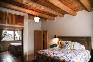 A bedroom with a wooden ceiling, a bed with patterned bedding, a nightstand with a lamp, and a window with curtains. - Peaceful Adobe home between Taos/TSV- Mountain Views, Hot Tub, Summer Garden (Valdez)