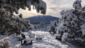 Uma área externa coberta de neve com bancos e mesas, rodeada por árvores carregadas de neve e montanhas ao fundo.